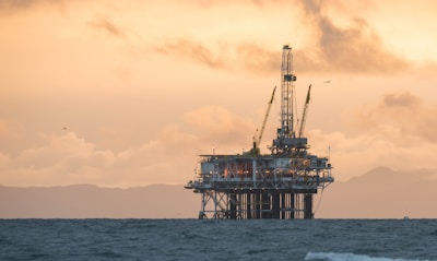 An offshore oil rig stands in the ocean against a backdrop of distant mountains and a dramatic sky, with a warm glow and scattered clouds.