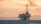 An offshore oil rig stands in the ocean against a backdrop of distant mountains and a dramatic sky, with a warm glow and scattered clouds.
