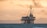 An offshore oil rig stands in the ocean against a backdrop of distant mountains and a dramatic sky, with a warm glow and scattered clouds.