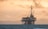 An offshore oil rig stands in the ocean against a backdrop of distant mountains and a dramatic sky, with a warm glow and scattered clouds.