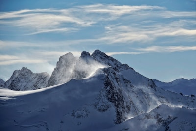 Snow-covered mountain peaks with a backdrop of wispy clouds. The crisp, cold atmosphere is accentuated by a light wind blowing snow off the ridges.