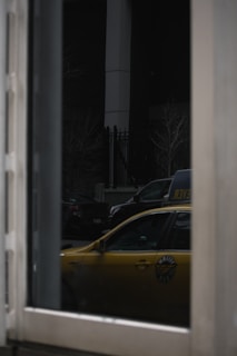 A reflection of a yellow taxi is visible in a window. The scene includes dark surroundings with parked cars and a building structure in the background. The reflection reveals part of the taxi's side and window.