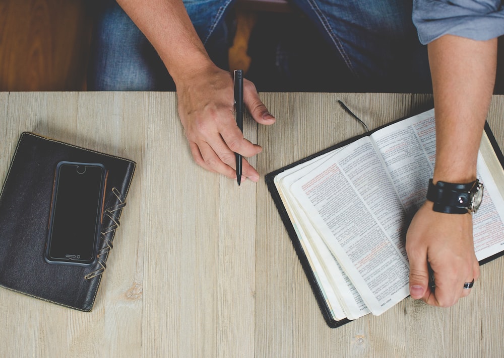 Man's hands holding an open book and a pen on a wooden surface with a notebook and a phone