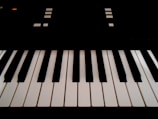 The image features a close-up view of piano keys, with alternating black and white keys in an orderly arrangement. There are geometric shapes and small lights visible in the background, suggesting parts of an electronic keyboard.
