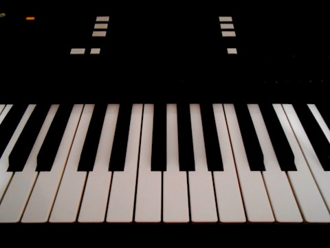 The image features a close-up view of piano keys, with alternating black and white keys in an orderly arrangement. There are geometric shapes and small lights visible in the background, suggesting parts of an electronic keyboard.