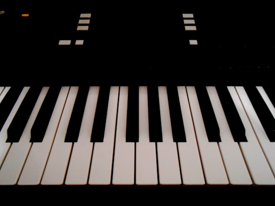 The image features a close-up view of piano keys, with alternating black and white keys in an orderly arrangement. There are geometric shapes and small lights visible in the background, suggesting parts of an electronic keyboard.