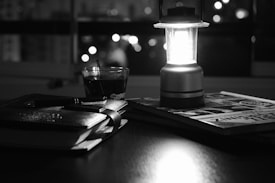 A dimly lit scene indoors with a bright lantern casting light on a stack of books and a notebook on a desk. A glass cup and a pen are also visible. The background is blurred, showing lights from outside the window, suggesting a nighttime setting.