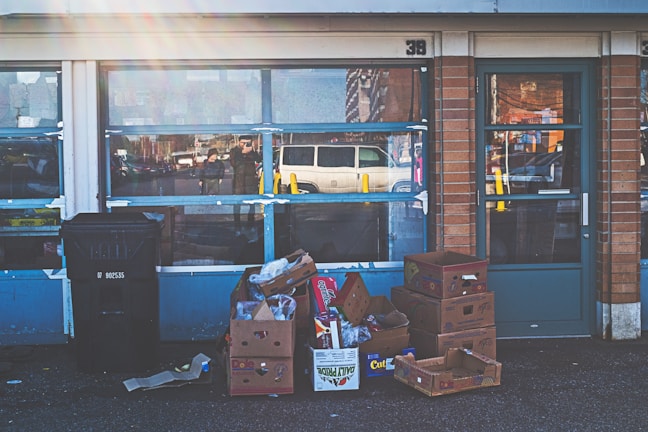 A storefront with a glass window reflecting a street scene, including a white van. Several cardboard boxes, some empty and others filled with miscellaneous items, are stacked on the ground outside the front, alongside a large black trash bin. The building features a combination of brickwork and blue paint.
