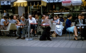 A lively outdoor café scene featuring people seated at tables, engaging in conversation and enjoying their time. Some individuals are reading newspapers while others are having drinks. A black poodle is sitting on the ground among the patrons. The background shows a counter with pastries and the café is covered with colorful canopies providing shade.