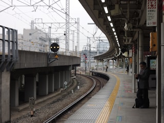 A train station platform with tracks curving to the left, surrounded by overhead wiring and modern urban infrastructure. A well-dressed man stands on the platform, looking at his phone. The platform is mostly empty except for a few distant figures. The setting is overcast, giving a subdued ambiance.