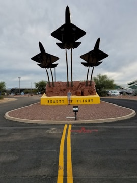 Three model aircraft on tall stands resembling flight takeoff, mounted on a reddish-brown rocky base with a yellow sign reading 'Beauty of Flight'. The display is set on a circular patch of gravel surrounded by road, with visible sky in the background.