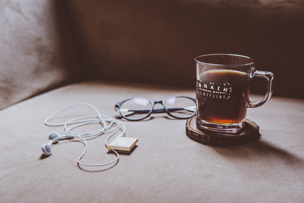 klarer Glasbecher mit Brain-Print auf braunem Holzplattenuntersetzer neben schwarz gerahmter Brille und weißen Kopfhörern