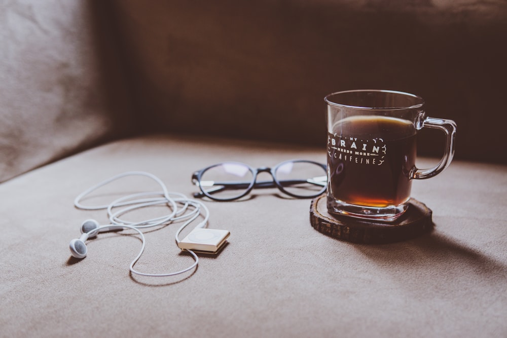 黒いフレームの眼鏡と白いイヤホンの横の茶色の木製スラブコースターにクリアブレインプリントガラスマグカップ