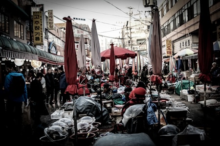 A bustling outdoor market scene with numerous closed red and beige umbrellas interspersed among stalls. Various goods are laid out, while people navigate the cramped aisles. Signage with Korean characters is visible on the surrounding buildings.