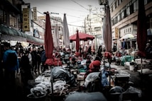A bustling outdoor market scene with numerous closed red and beige umbrellas interspersed among stalls. Various goods are laid out, while people navigate the cramped aisles. Signage with Korean characters is visible on the surrounding buildings.