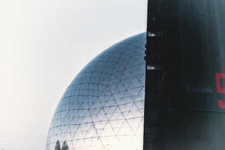 A large geodesic dome with a reflective surface partially obscured by a dark, rectangular structure on the right. The dome's framework is visible, consisting of a network of interconnected triangles, and is set against a pale sky. A partial number is in red on the right structure.