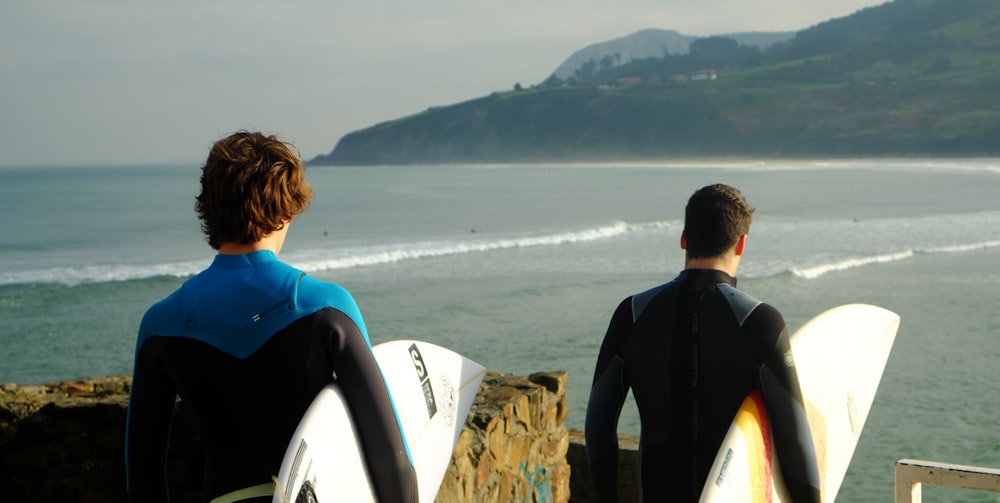 two male wearing wetsuits holding surfboards standing nearby each other on top of hill with view of beach below