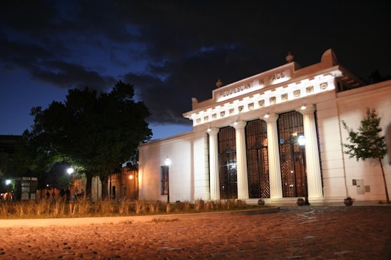 A well-lit neoclassical-style building at night with the text 'REQUESCANT IN PACE' inscribed above the entrance. The structure features tall columns and a walkway lined with low shrubs and grass. The surrounding area includes cobblestone pavement and large trees, under a partially clouded night sky.