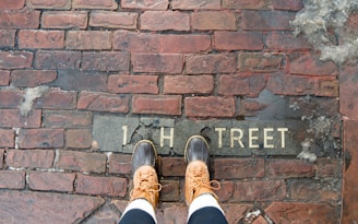 A pair of feet wearing brown leather boots standing on a wet brick pavement. The pavement has the partially visible text '1 H TREET' engraved on it.