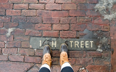 A pair of feet wearing brown leather boots standing on a wet brick pavement. The pavement has the partially visible text '1 H TREET' engraved on it.