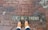 A pair of feet wearing brown leather boots standing on a wet brick pavement. The pavement has the partially visible text '1 H TREET' engraved on it.