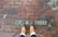 A pair of feet wearing brown leather boots standing on a wet brick pavement. The pavement has the partially visible text '1 H TREET' engraved on it.