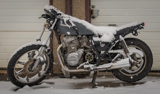 A motorcycle is parked outdoors, covered in a layer of snow, which highlights its frame and handlebars. The bike rests on a kickstand in front of a closed garage door, with visible branding on its body.