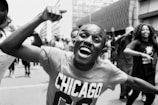 A joyful crowd of people participating in a public event on a city street. The central figure is a man with an enthusiastic expression, raising his arm. He wears a T-shirt with 'Chicago' written on it. Other participants in the background are smiling and moving energetically.