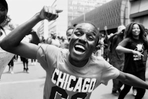 A joyful crowd of people participating in a public event on a city street. The central figure is a man with an enthusiastic expression, raising his arm. He wears a T-shirt with 'Chicago' written on it. Other participants in the background are smiling and moving energetically.