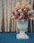 A large decorative vase filled with a variety of artificial flowers, including orange, white, and beige blooms, is set against a golden curtain backdrop. The vase features a classic design and rests on a blue carpeted floor.