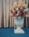 A large decorative vase filled with a variety of artificial flowers, including orange, white, and beige blooms, is set against a golden curtain backdrop. The vase features a classic design and rests on a blue carpeted floor.