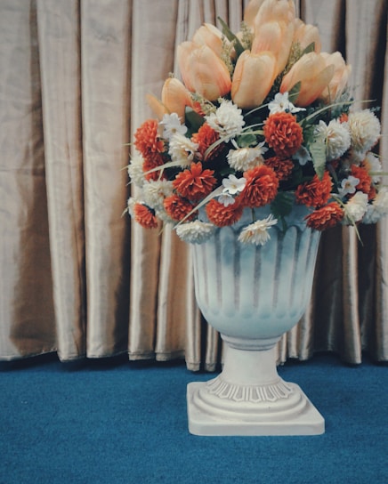 A large decorative vase filled with a variety of artificial flowers, including orange, white, and beige blooms, is set against a golden curtain backdrop. The vase features a classic design and rests on a blue carpeted floor.