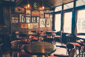 A cozy interior of a pub with a wooden theme. Multiple round wooden tables and chairs are arranged neatly across the room. The walls are adorned with vintage posters and signs, creating a nostalgic ambiance. Soft, warm lighting illuminates the space, enhancing the vintage décor. Large windows with curtains allow natural light to filter through.