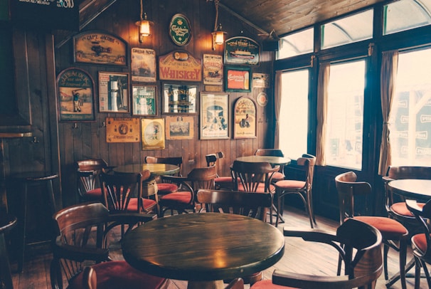 A cozy interior of a pub with a wooden theme. Multiple round wooden tables and chairs are arranged neatly across the room. The walls are adorned with vintage posters and signs, creating a nostalgic ambiance. Soft, warm lighting illuminates the space, enhancing the vintage décor. Large windows with curtains allow natural light to filter through.