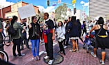 A group of people gathers on a city street, engaging in conversation and holding signs. One sign reads 'Imagine Peace'. The setting appears to be a demonstration or rally, with participants of diverse backgrounds. Buildings and trees are visible in the background under a partly cloudy sky.