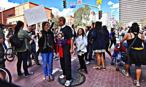 A group of people gathers on a city street, engaging in conversation and holding signs. One sign reads 'Imagine Peace'. The setting appears to be a demonstration or rally, with participants of diverse backgrounds. Buildings and trees are visible in the background under a partly cloudy sky.