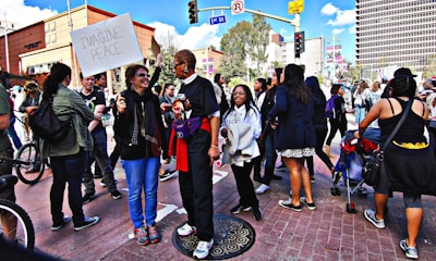 A group of people gathers on a city street, engaging in conversation and holding signs. One sign reads 'Imagine Peace'. The setting appears to be a demonstration or rally, with participants of diverse backgrounds. Buildings and trees are visible in the background under a partly cloudy sky.