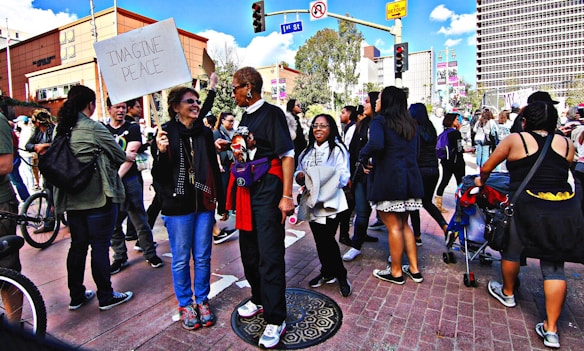 A group of people gathers on a city street, engaging in conversation and holding signs. One sign reads 'Imagine Peace'. The setting appears to be a demonstration or rally, with participants of diverse backgrounds. Buildings and trees are visible in the background under a partly cloudy sky.