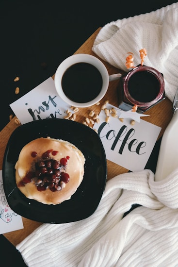 A cozy breakfast scene with a cup of black coffee and a stack of pancakes topped with berries and syrup on a black plate. Beside the coffee, a jar of jam and scattered nuts add to the setting. Artistic cards with calligraphy words 'just have coffee' are placed nearby, along with a soft white knit scarf, creating a warm and inviting atmosphere.