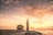 A church with a tall bell tower stands on a rocky shoreline, overlooking a tranquil sea at sunset. The sky is filled with soft clouds illuminated by warm, golden light, creating a serene atmosphere.