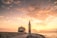 A church with a tall bell tower stands on a rocky shoreline, overlooking a tranquil sea at sunset. The sky is filled with soft clouds illuminated by warm, golden light, creating a serene atmosphere.