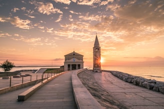 A serene coastal scene at sunset, featuring a small church with a bell tower on the right. Sunlight beautifully reflects off the calm sea to the left, and a pathway lined with a railing leads toward the church. The sky is filled with scattered clouds tinted with soft hues of pink and orange.