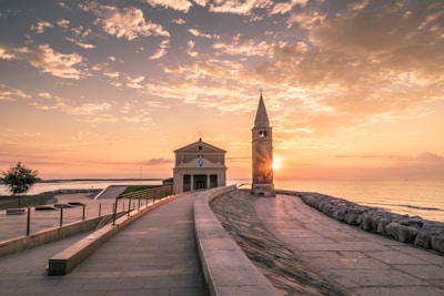 A serene coastal scene at sunset, featuring a small church with a bell tower on the right. Sunlight beautifully reflects off the calm sea to the left, and a pathway lined with a railing leads toward the church. The sky is filled with scattered clouds tinted with soft hues of pink and orange.