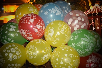 Eleven colorful balloons with intricate white designs and patterns are clustered together. The balloons include red, yellow, blue, and green colors, adorned with floral, animal, and abstract patterns. A warm, ambient light in the background suggests a festive atmosphere, possibly taken during an evening celebration or event.