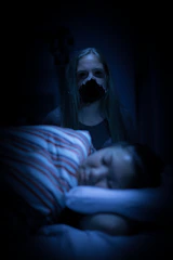 A dimly lit scene features a young person sleeping peacefully in the foreground, with their head resting on a pillow. A figure with long hair stands ominously in the background, their face partially obscured by darkness, contributing to an unsettling atmosphere.