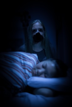 A dimly lit scene features a young person sleeping peacefully in the foreground, with their head resting on a pillow. A figure with long hair stands ominously in the background, their face partially obscured by darkness, contributing to an unsettling atmosphere.