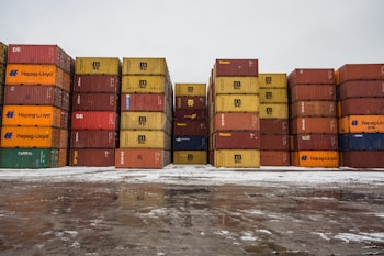 Stacks of colorful shipping containers are arranged in an outdoor yard. The containers are of various brands like MSC and Hapag-Lloyd, and they are stacked on top of a snowy and wet surface under a cloudy sky.