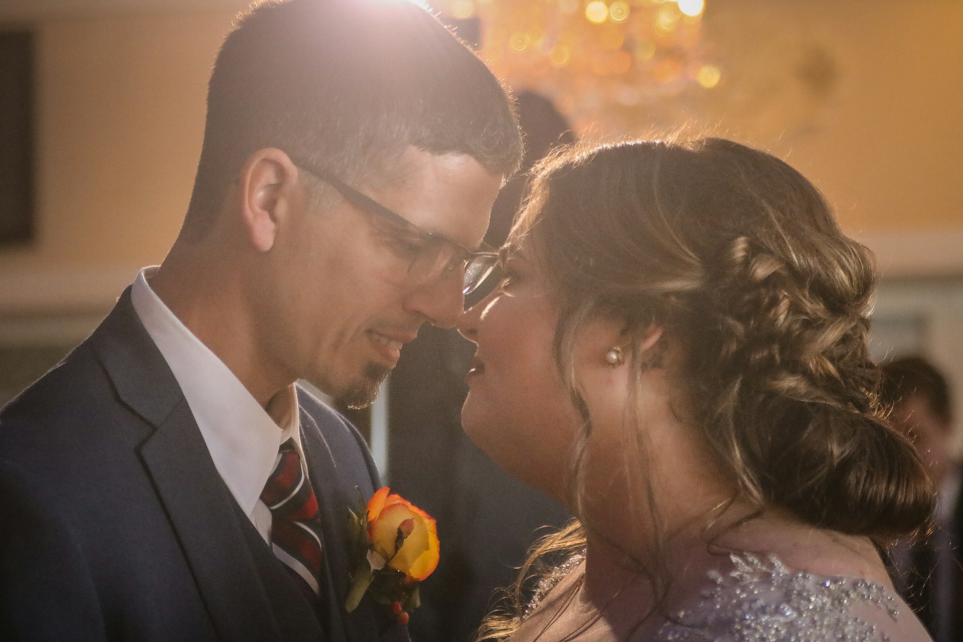 A couple is shown in a close, intimate moment, possibly during a dance. The man is wearing glasses, a suit, and a tie adorned with a floral boutonniere. The woman, with her hair styled in an elegant updo, is wearing a dress with sparkling embellishments. Warm, soft lighting illuminates the scene, creating a romantic atmosphere.