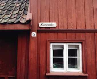 A rustic red wooden building with a small, white-framed window. Above the window, there is a sign with the text 'Damstredet.' Next to this sign is a plaque with the number 5. The roof has dark-colored tiles with patches of green moss or weathering.