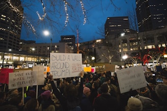 A large group of people gathered in an urban area during the evening, holding various protest signs. The signs feature messages related to political dissent, some specifically targeting policies associated with Donald Trump. Buildings and trees decorated with lights form the backdrop, and the setting appears to be in a city square.